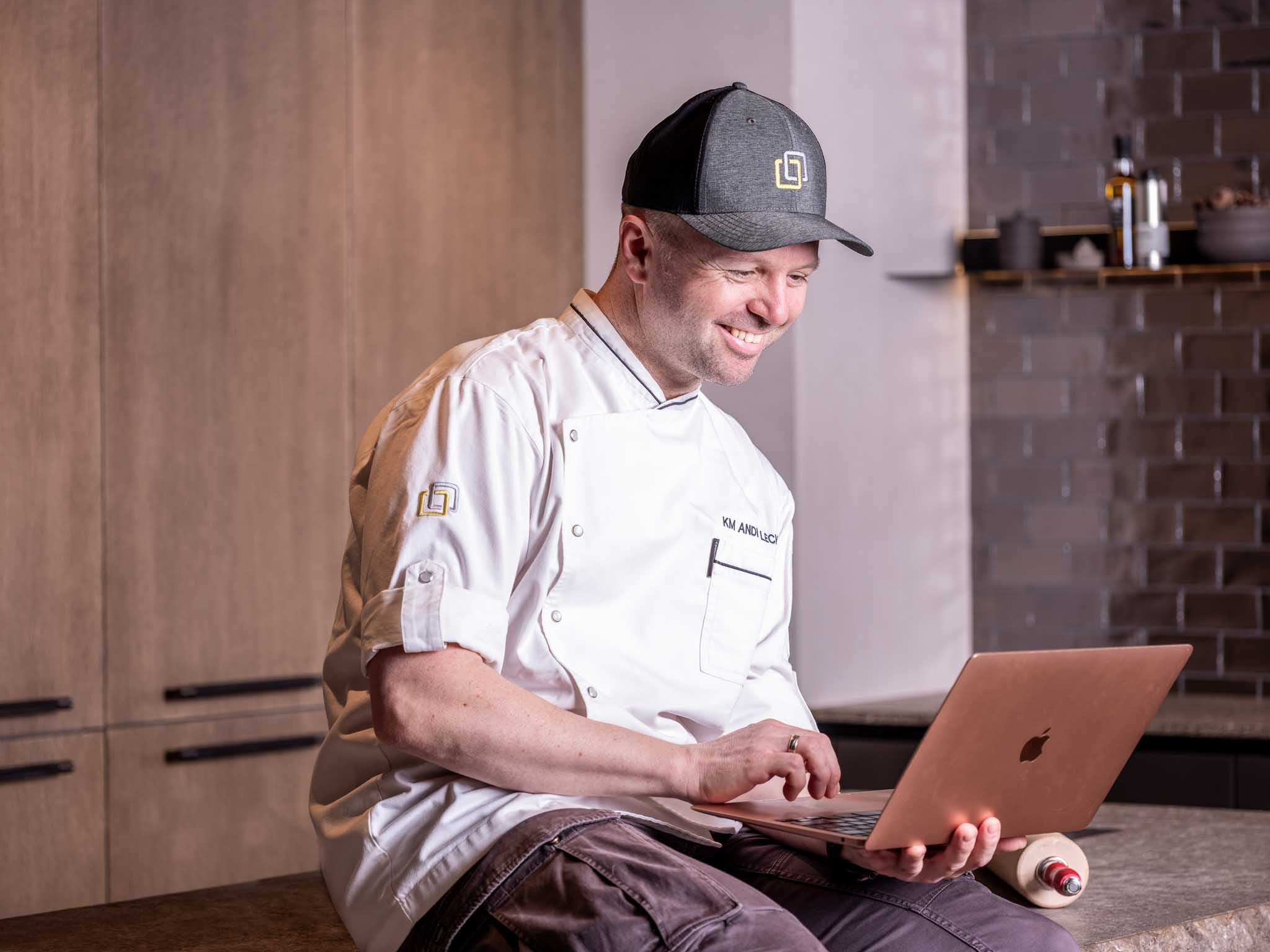 AndreasLechner_Laptop Ein Foto von Geschäftsführer Andreas Lechner, grinsend sitzend auf einer Küchenanrichte mit einem Laptop in der Hand