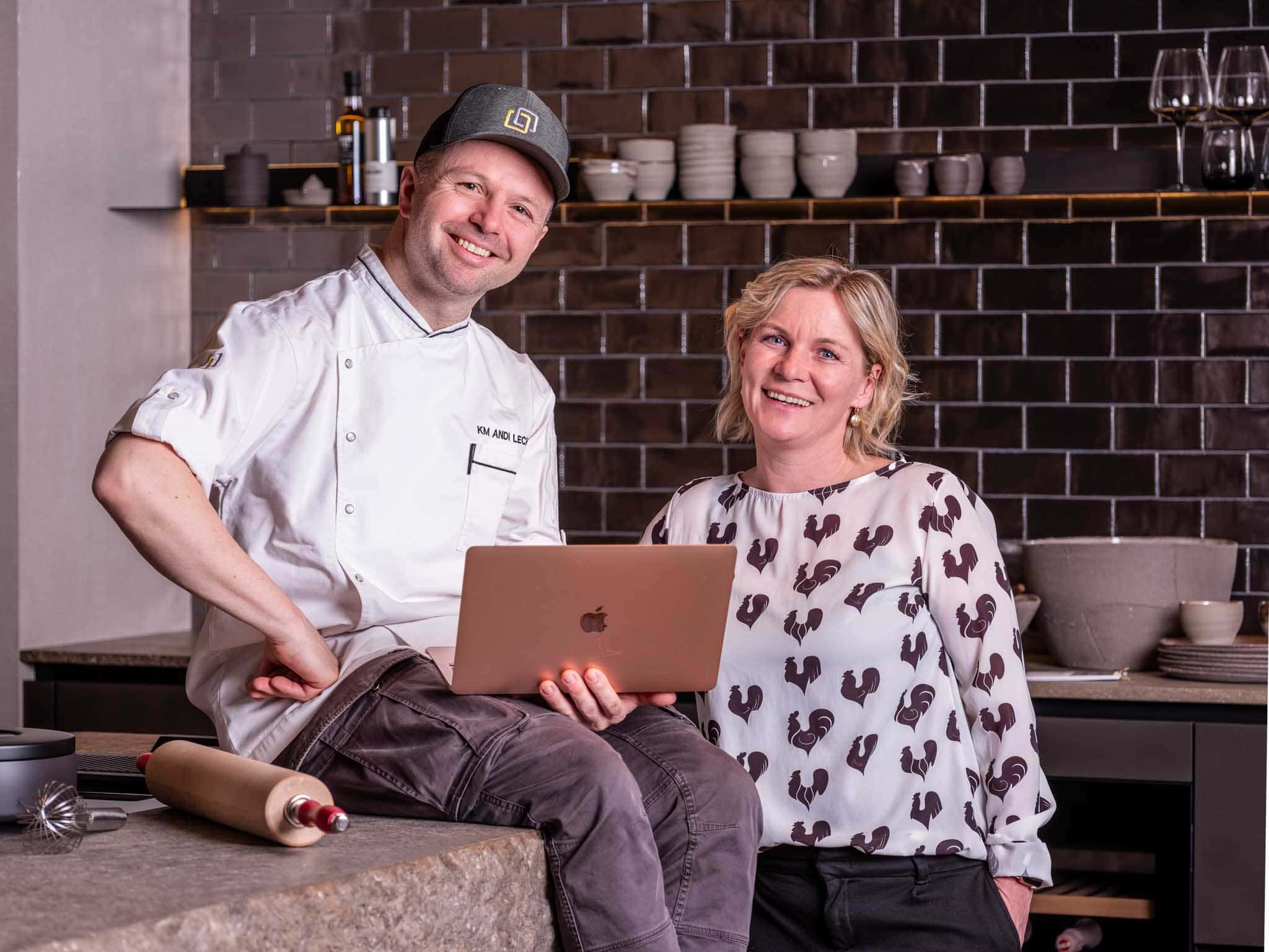 Andi und Bianca Lechner Ein Foto von Andreas lechner grinsend sitzend auf einer Küchenanrichte mit einem Lap in der Hand. Neben ihm steht seine Frau Bianca Lechner.