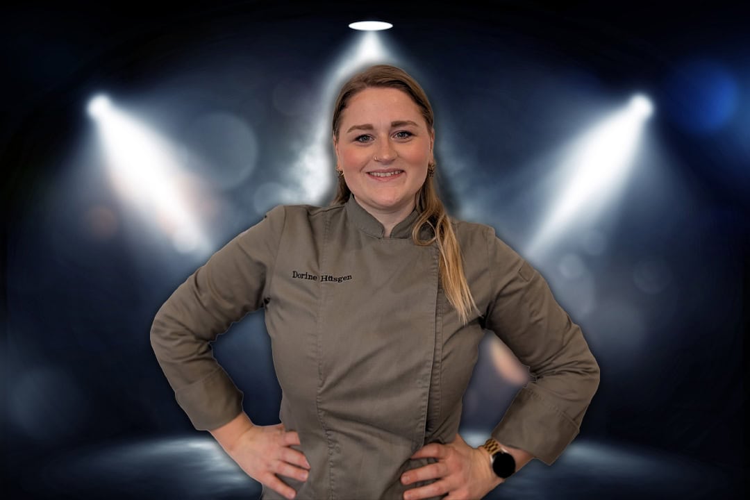 Portrait Foto von der Spitzenköchin Dorine Hüsgen mit einer braunen Küchenjacke grinsend vor dunkelblauem Hintergrund